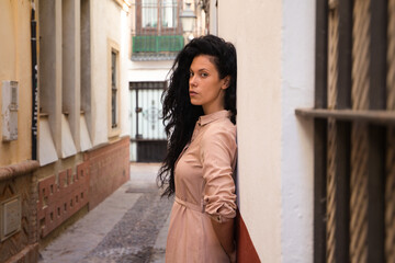 beautiful woman with dark curly hair leaning against the wall of a typical european house in a mediterranean style city. The woman is serious and sad. Concept expressions.