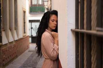 beautiful woman with dark curly hair leaning against the wall of a typical european house in a mediterranean style city. The woman is serious and sad. Concept expressions.