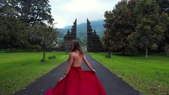 Pretty asian woman walks to balinese gates in long red dress and drone follows in slowmotion on Hangara gates in Bali Indonesia. fashion concept. High quality fullhd aerial footage