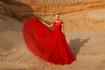 Full length image of a young woman in red waving dress with flying fabric on the background of golden sands.