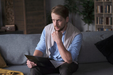 Confident European business male working remotely use laptop on couch at cozy living room. Modern...