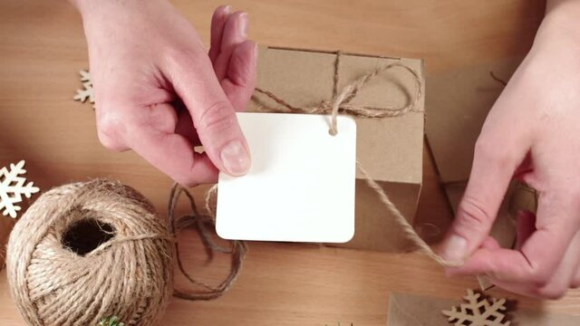 Eco-friendly gifts, Homemade gifts, DIY. Hands attaching a blank label to a kraft paper box with a twine bow on a table with snowflakes, Full HD.