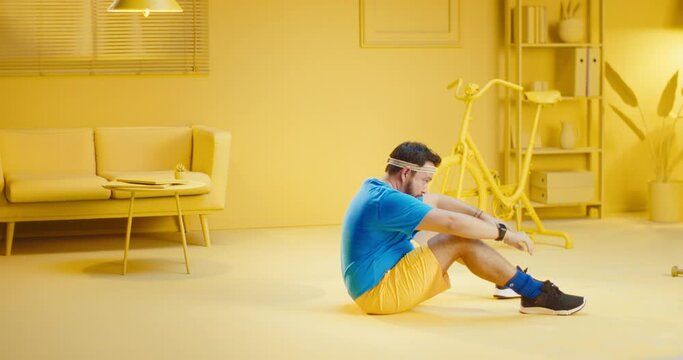 Exhausted overweight bearded young man doing sit ups and taking a break while lying on the floor at home with monochrome yellow interior. Full length shot of a fat male model doing sit up exercise