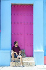 Front view of man with backpack sit on beautiful pink door in Cartagena de Indias. Vertical view of male tourist resting on house with pink and blue walls. Travel to Colombia concept. - obrazy, fototapety, plakaty
