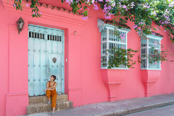 Side view of woman sit on a pink wall in Cartagena de Indias. Horizontal view of latin woman sightseeing in spanish historic ancient city. Travel to Colombia concept.