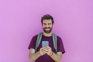 front view of caucasian man holding a phone on pink background. Horizontal view of latin american man using technology isolated on pink wall. People and travel concept.