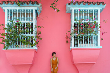 Front view of woman standing on a pink wall with balcony in Cartagena de Indias. Horizontal view of latin woman sightseeing in spanish historic ancient city. Travel to Colombia concept. - obrazy, fototapety, plakaty