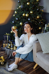 A beautiful girl is sitting at the Christmas tree and holding a Christmas tree decoration. New Year's mood. Merry Christmas.