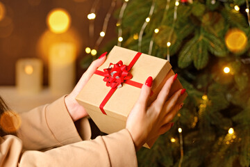 Female hands holding craft gift box with red ribbon near Christmas tree decorated with lights
