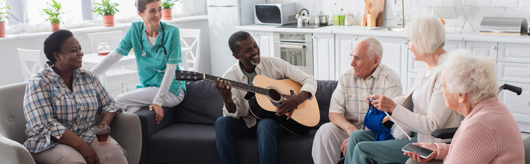 Smiling interracial elderly people playing acoustic guitar near nurse in nursing home, banner