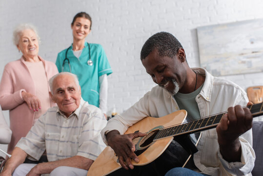 African american pensioner playing acoustic guitar near friends and nurse in nursing home