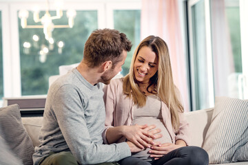 Young pregnant happy couple sitting on couch husband stroking baby belly of blond wife