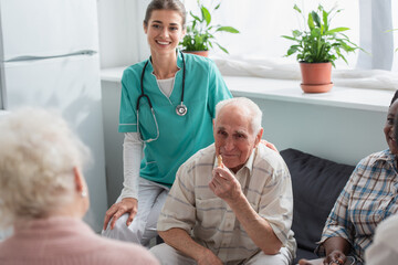 Smiling nurse sitting near senior interracial patients with chess figure in nursing home
