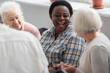 Excited african american woman looking at senior friend with tea in nursing home
