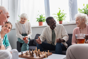 Cheerful multiethnic people with tea playing chess near nurse in nursing home