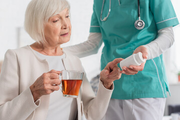 Nurse holding pills near senior patient with tea in nursing home