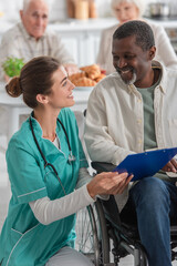 Obraz premium Cheerful nurse holding clipboard near african american patient in wheelchair in nursing home