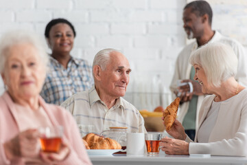 Smiling senior woman holding croissant and tea near friend in nursing home
