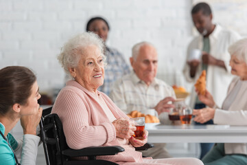 Senior woman holding tea in wheelchair near nurse and blurred interracial friends in nursing home