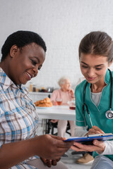 Smiling nurse and african american patient looking at clipboard in nursing home