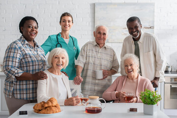 Smiling nurse standing near interracial patients and tea in nursing home
