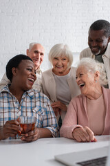 Fototapeta premium African american woman holding tea near smiling friends and laptop in nursing home