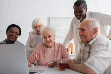 Smiling senior woman holding smartphone and pointing at laptop near interracial friends in nursing home