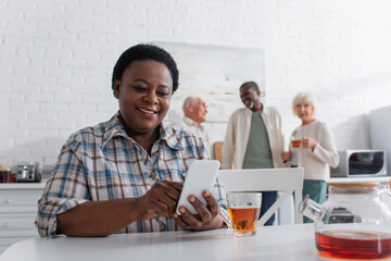 Smiling african american woman using smartphone near tea and blurred people in nursing home
