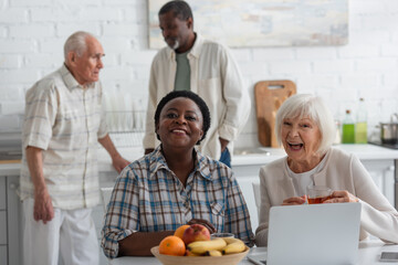 Cheerful multiethnic women with tea looking at camera near devices and fruits in nursing home