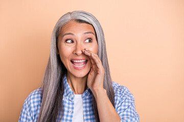 Photo of cute excited retired woman wear plaid shirt arm lips making announcement looking empty space isolated beige color background