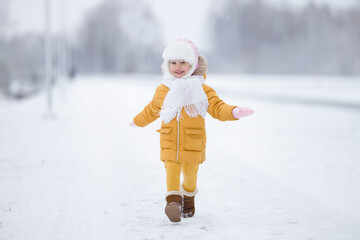 One happy smiling baby girl in warm clothes running on white snow covered road in cold winter day. Front view. 2 years old toddler.