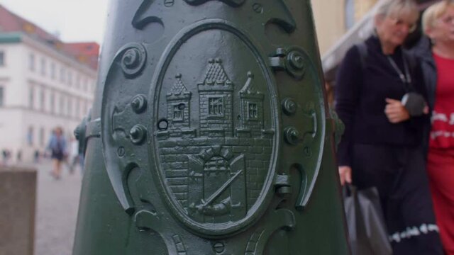 Coat of arms of the city of Prague on a lamppost. Passers-by move in bokeh. Visual identification mark (a subspecies of emblems), compiled and used according to the rules of heraldry.