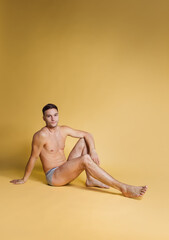 One young handsome shirtless brown-eyed man sitting on floor isolated on yellow studio background....