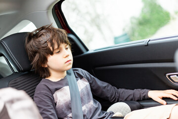 Sad pensive boy looking down is sitting in a car wearing a seat belt. Unhappy child travelling in...
