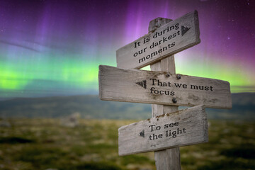 it is during our darkest moments text quote on signpost under the northern lights.