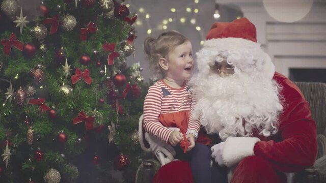 Santa Claus talking to cute toddler girl in decorated living room. Real time
