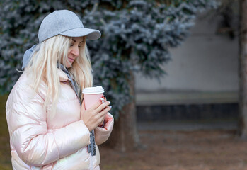 Attractive young woman girl in a pink jacket and a cap stands in the park with a cup of tea and coffee in her hands, selective focus, copy space for text