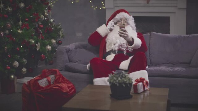 Man in Santa Claus costume taking selfie on smartphone sitting on couch