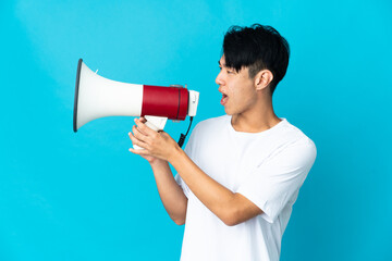 Young Chinese man isolated on blue background shouting through a megaphone to announce something in...
