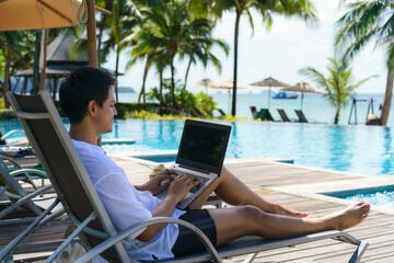 Asian man spent his summer vacation working on his laptop in a chair near the swimming pool in resort hotel near sea.. - obrazy, fototapety, plakaty