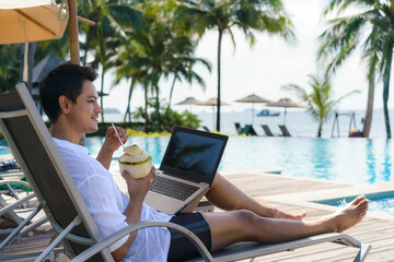 Asian man drinking coconut water while he is working on his laptop on a chairnear pool at resort...