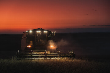 Combine harvester working in a wheat field at night with lights on and red sky at sunset