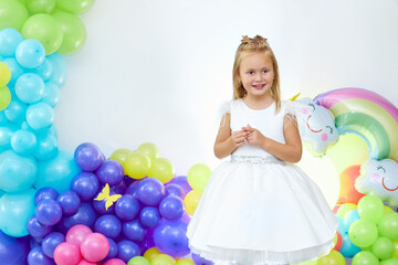 Fototapeta na wymiar A beautiful blonde girl five years old in a white dress stands on the background of balloons. Portrait of a funny child. Children's birthday concept.