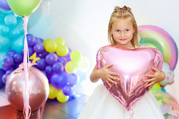 Fototapeta na wymiar A beautiful blonde girl of five years old is holding a heart-shaped balloon. Portrait of a funny child. Children's birthday concept.