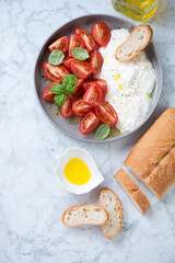 Salad with red cherry tomatoes and italian stracciatella cheese, top view on a light-grey marble background, vertical shot