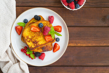 A stack of French toast on a plate with fresh berries, almond petals and honey on a dark wooden background. Delicious breakfast. Copy space.