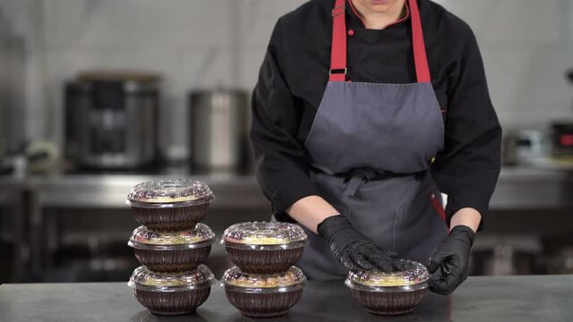 in a restaurant kitchen, a woman piles up prepackaged meals with healthy food in a plastic package on a metal table in a food delivery service, copy space.