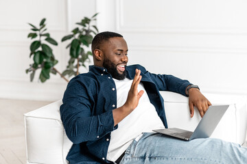 Young African-American freelancer guy sitting with the laptop during video meeting, working on a distance, talking with potential clients and partners, looking at the laptop screen and waving hello