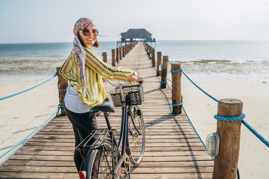 Woman looking at camera and smiling dressed in light summer clothes, sunglasses with bicycle on the wooden sea pier on sandy Zanzibar beach.Careless vacation in the tropical countries concept image