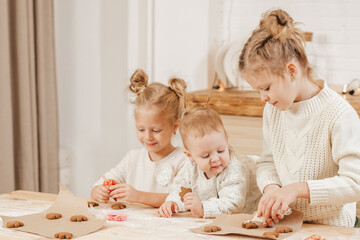 3 blonde girls decorate Christmas cookies with icing on a wooden table in the kitchen. The girlfriends are cooking together in the kitchen with Christmas decor. Warm relationship of sisters. 